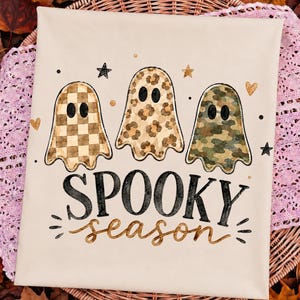 May include: A cream-colored t-shirt featuring a graphic of three ghosts with black eyes. The left ghost has a tan checkerboard pattern, the middle has a leopard print, and the right has a camouflage pattern. The text SPOOKY season is printed below.