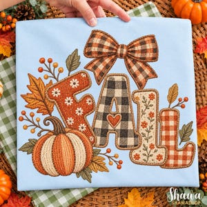 May include: A light blue t-shirt featuring an embroidered design of the word FALL. The letters are decorated with orange floral patterns, black and white buffalo check, and a brown plaid bow. A large orange pumpkin sits at the base.