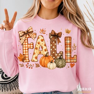 May include: A light pink crewneck sweatshirt featuring the word FALL in large decorative letters. The letters are patterned with orange plaid, autumn leaves, and brown polka dots, accented by two brown bows and three pumpkins in orange, white, and green.