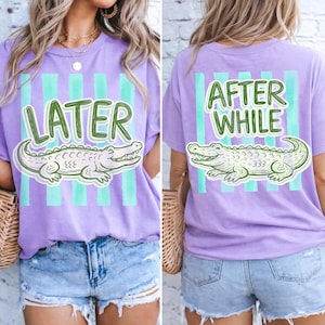 May include: A lilac-colored t-shirt with a green and white alligator graphic. The front of the shirt says "LATER" in green, and the back says "AFTER WHILE". The shirt has vertical teal stripes.