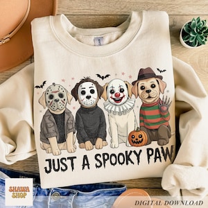 May include: A cream-colored crewneck sweatshirt featuring a graphic of four dogs dressed in horror movie costumes. The text "JUST A SPOOKY PAW" is printed below the dogs. The illustration includes bats, stars, and a small pumpkin.