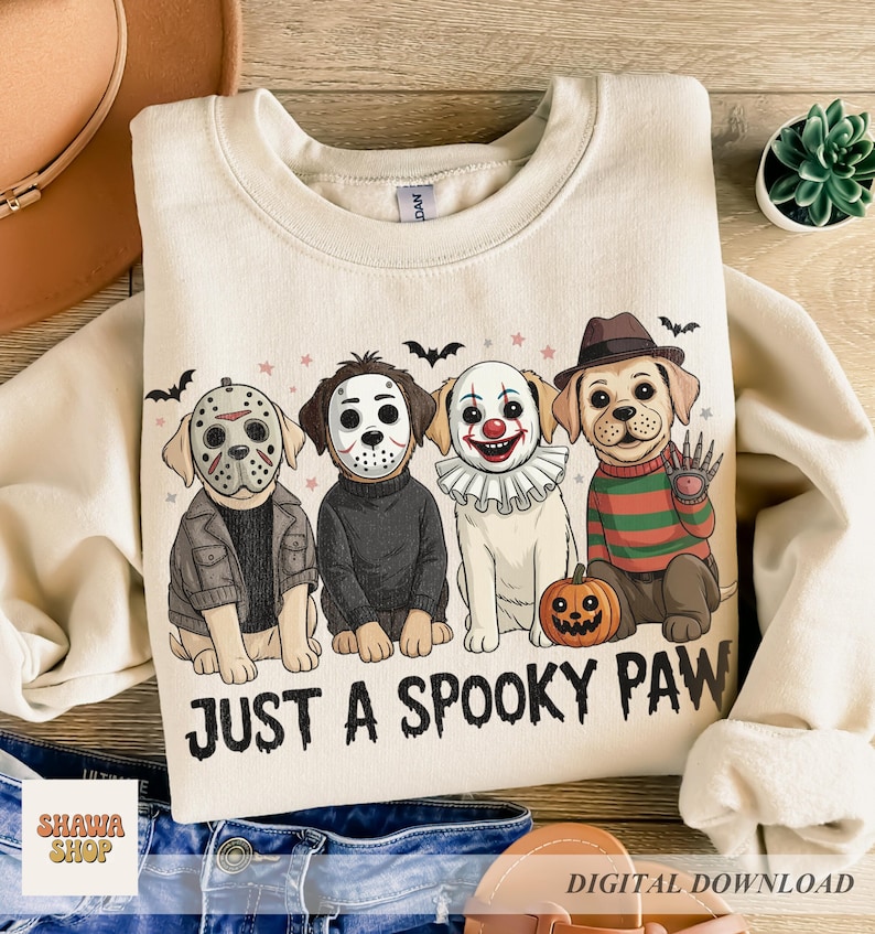 May include: A cream-colored crewneck sweatshirt featuring a graphic of four dogs dressed in horror movie costumes. The text "JUST A SPOOKY PAW" is printed below the dogs. The illustration includes bats, stars, and a small pumpkin.