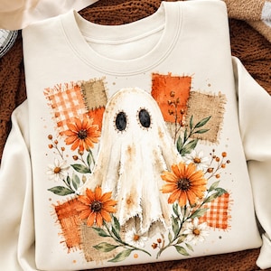 May include: A cream-coloured crewneck sweatshirt featuring a central graphic of a ghost with black stitched eyes. The design includes orange and tan plaid fabric patches, vibrant orange sunflowers, white daisies, and green leafy stems.
