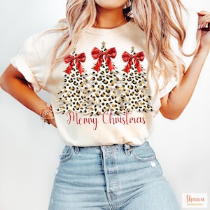 May include: A white t-shirt with a graphic of three leopard print Christmas trees with red bows and gold stars. The text "Merry Christmas" is printed below the trees.