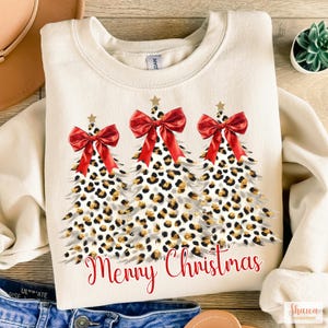 May include: A cream-colored sweatshirt with a festive design featuring three leopard print Christmas trees with red bows and gold glitter. The text "Merry Christmas" is printed in red below the trees.