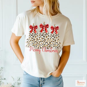 May include: White t-shirt with a graphic of three leopard print Christmas trees with red bows and gold stars. The text "Merry Christmas" is printed below the trees.