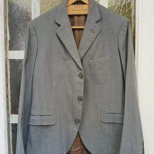 May include: A gray blazer with a notched lapel, three buttons, and two front pockets. The blazer is hanging on a wooden hanger. The interior lining is brown. The hanger has the text "ETON Sartoria de Moda".