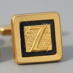 Może przedstawiać: Złote kwadratowe spinki do mankietów z czarnym środkiem. Środek przedstawia literę "Z" w kolorze złotym z ozdobnym wzorem. Spinka do mankietu ma teksturowaną obwódkę i błyszczące wykończenie, do formalnych strojów.