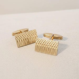 Puede incluir: Un par de gemelos dorados con un diseño geométrico texturizado. La cara rectangular de cada gemelo presenta un patrón repetido de pequeñas formas en relieve. Los gemelos se muestran sobre una superficie de color claro.