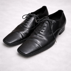 Puede incluir: Un par de zapatos de vestir de cuero negro con puntera cuadrada y cierre de cordones. Los zapatos tienen un diseño clásico, adecuado para ocasiones formales. El cuero parece liso y las costuras son visibles alrededor de las suelas.