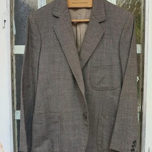 Puede incluir: Una blazer de tweed marrón con cierre de dos botones y dos bolsillos delanteros. La blazer está colgada de una percha de madera. La blazer tiene un diseño clásico y a medida.