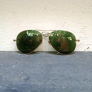 Puede incluir: Gafas de sol de aviador con montura de metal dorado y lentes verdes. Las lentes reflejan el entorno. Las gafas de sol están sobre una superficie gris oscuro.