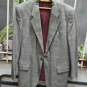Puede incluir: Una blazer gris con un patrón texturizado, con forro burdeos. La blazer tiene cierre de dos botones, solapas con muescas y dos bolsillos delanteros. Está colgada en una percha de madera.