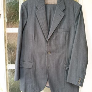 Puede incluir: Un traje gris, compuesto por una chaqueta y un pantalón, cuelga de una percha de madera. La chaqueta tiene cierre de dos botones, solapas con muescas y tres botones en cada puño. El traje está hecho de una tela texturizada con un sutil patrón de rayas verticales.