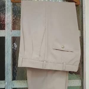 Puede incluir: Un pantalón de color beige claro con un bolsillo de ojal con cierre de botón en la parte delantera, cintura con trabillas para cinturón y raya central, colgado de una percha de madera con la inscripción Casa Martinez H. Irigoyen 982.