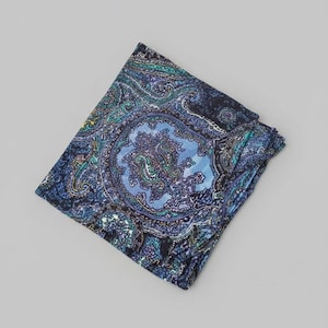 Puede incluir: Pañuelo de bolsillo plegado con un estampado de cachemira en tonos azules, turquesas y morados. El intrincado diseño presenta un medallón central y bordes detallados, creando un accesorio clásico.