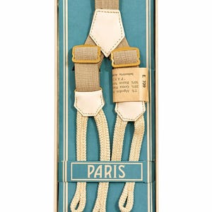 Puede incluir: Tirantes color beige con detalles de cuero crema y herrajes metálicos dorados, presentados en una caja azul. La palabra "PARIS" está impresa en la caja. Los tirantes están hechos de 7% algodón, 25% goma y 10% rayón.