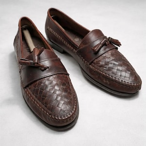 May include: A pair of brown leather loafers. The shoes have woven detailing on the toe and a tassel accent. The classic design and dark brown colour make them suitable for various occasions.