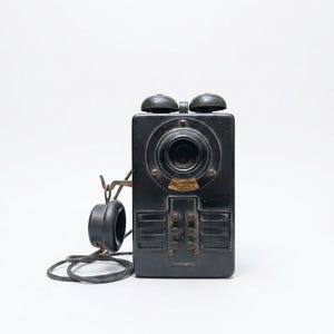 May include: An antique black telephone with a rectangular body, two dome-shaped bells, and a circular earpiece. The phone has a dial and several buttons, with a cord connecting the earpiece to the main unit. A vintage communication device.