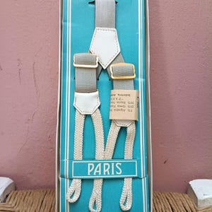 May include: Gray suspenders with white leather accents and gold-colored metal hardware, displayed in a blue box. The box has the word "PARIS" printed on it. The suspenders are made of a blend of rayon, cotton, and rubber.