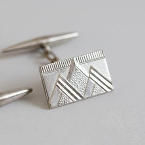 Puede incluir: Gemelos plateados con un diseño geométrico. Los gemelos rectangulares presentan un patrón texturizado con rayas diagonales y un centro texturizado. El poste del gemelo es visible en el fondo.