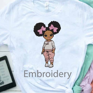 May include: Embroidered design of a girl with brown skin, black hair in two puffs with pink bows, wearing a pink and white striped shirt and pink pants. The design is on a light blue t-shirt with the word "Embroidery" printed below.