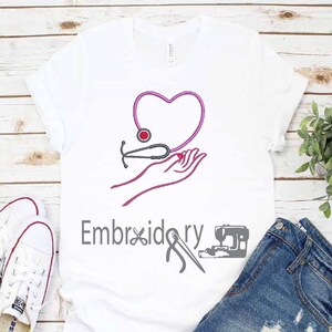 Puede incluir: Camiseta blanca con bordado rosa de un corazón, un estetoscopio y manos. El texto "Bordado" también está bordado en gris en la camiseta.