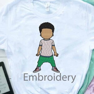 May include: A light blue t-shirt with an embroidered design of a cartoon boy wearing a green shirt and pants. The text "Embroidery" is embroidered in gray below the boy.
