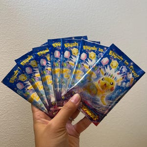 May include: A fan of six blue rectangular trading card booster packs featuring a yellow Pikachu character wearing a crown, surrounded by colorful gems and the text Elijah is five and Raging Sparks printed on the front of each individual pack.