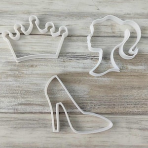 Pode incluir: Três cortadores de biscoitos de plástico branco em forma de coroa, sapato de salto alto e perfil de mulher.