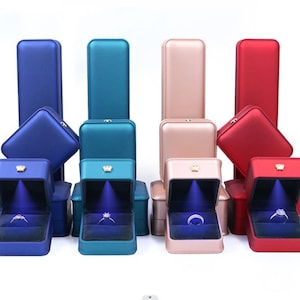 May include: Four rectangular jewelry boxes in blue, teal, rose gold, and red, each featuring a hinged lid and an integrated LED light that illuminates a ring placed inside on a dark velvet cushion. Each box has a small gold-tone crown emblem.
