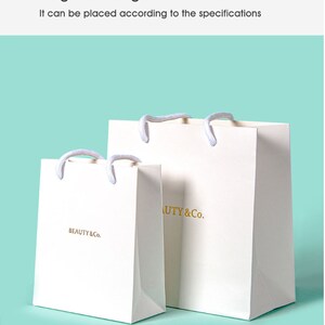 May include: Two white paper shopping bags with rope handles. The bags have the text "BEAUTY & Co." printed in gold. The bags are on a light blue background. The text "Large size bags" and "It can be placed according to the specifications" are at the top.