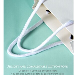 May include: Close-up of a cream-colored paper tote bag with white cotton rope handles. The bag has a textured surface and is set against a light teal background. Text on the image reads: "USE SOFT AND COMFORTABLE COTTON ROPE."