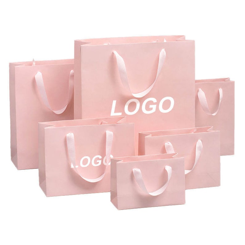 May include: A collection of pink paper shopping bags in various sizes. Each bag has a white satin ribbon handle and the word "LOGO" printed in white. The bags are arranged on a white background.