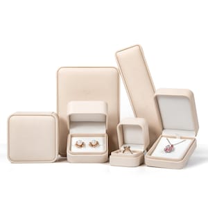 May include: A collection of beige jewelry boxes in various shapes and sizes. Some boxes are open, displaying a pair of pearl earrings, a ring, and a pendant necklace. The boxes have a soft, neutral color and a minimalist design.
