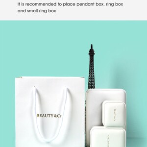 May include: A white paper bag with the text "BEAUTY & Co." in gold lettering, along with a small Eiffel Tower figurine and white jewelry boxes. The image is set against a light teal background.