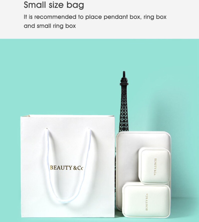 May include: A white paper bag with the text "BEAUTY & Co." in gold lettering, along with a small Eiffel Tower figurine and white jewelry boxes. The image is set against a light teal background.