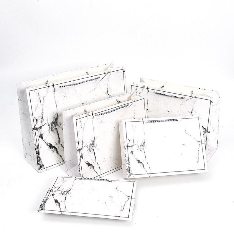May include: A collection of gift bags in various sizes, featuring a white marble design with black veining. Each bag has a black border and a matching rope handle. The bags are arranged on a white background.