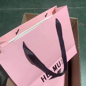 May include: A pink paper shopping bag with black handles. The bag has the text "HE MU" printed on the front. The bag is inside a cardboard box.