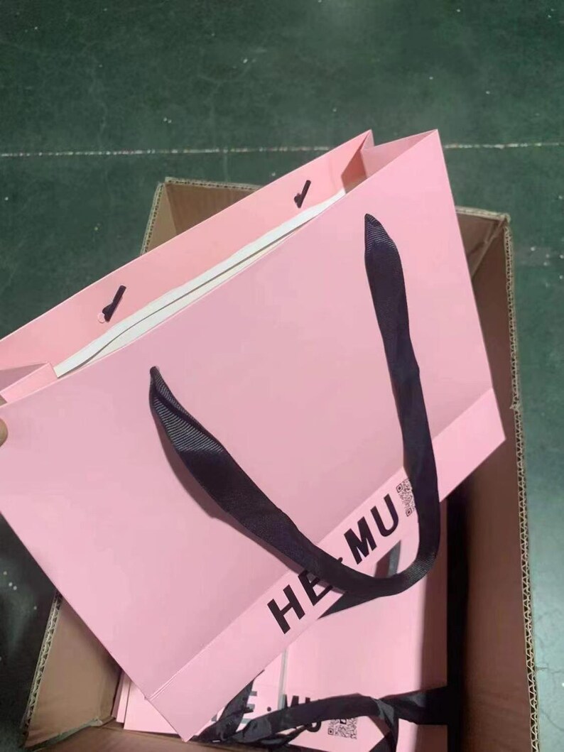 May include: A pink paper shopping bag with black handles. The bag has the text "HE MU" printed on the front. The bag is inside a cardboard box.