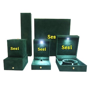 May include: A collection of dark green velvet jewelry boxes in various shapes and sizes. Each box has a gold-colored clasp and the word "Sesi" printed in yellow. Some boxes have interior lighting.