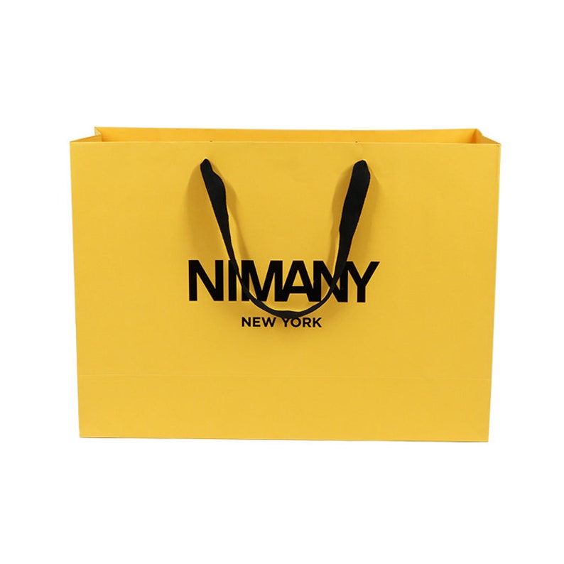 May include: A yellow shopping bag with black handles and the text "NIMANY NEW YORK" printed in black. The bag is rectangular and has a simple, clean design, suitable for retail or gift packaging.
