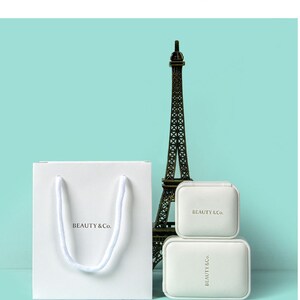 May include: A white paper shopping bag with a rope handle and the text "BEAUTY & Co." is next to two white jewelry boxes and a miniature Eiffel Tower. The boxes and bag are on a light blue background.