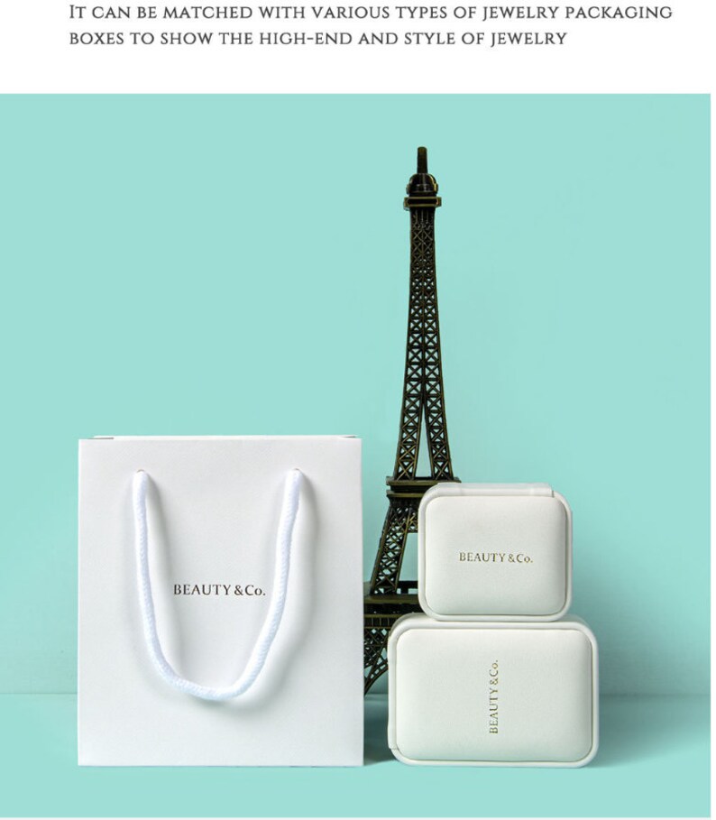 May include: A white paper shopping bag with a rope handle and the text "BEAUTY & Co." is next to two white jewelry boxes and a miniature Eiffel Tower. The boxes and bag are on a light blue background.