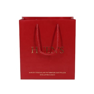 May include: A red shopping bag with gold lettering that reads "HUENS". The bag has red rope handles and additional gold text at the bottom. The text at the bottom reads "14 SCOTTS ROAD, #02-78/79/80 FAR EAST PLAZA SINGAPORE 228213".
