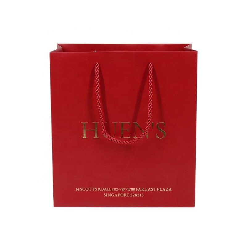May include: A red shopping bag with gold lettering that reads "HUENS". The bag has red rope handles and additional gold text at the bottom. The text at the bottom reads "14 SCOTTS ROAD, #02-78/79/80 FAR EAST PLAZA SINGAPORE 228213".