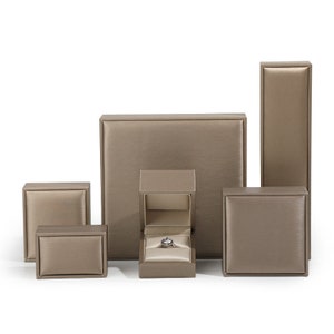May include: A collection of six metallic bronze jewelry display boxes of varying shapes and sizes, including one tall rectangular box, one large square box, and one open square box containing a silver ring with a clear center stone.