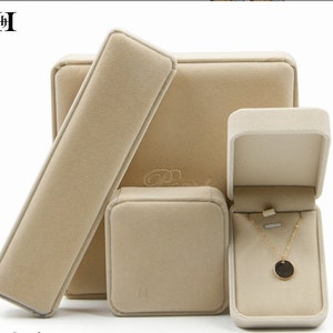 Puede incluir: Un conjunto de cajas de exhibición de joyas beige y un collar. Las cajas son de varias formas y tamaños, incluyendo una caja rectangular, una caja cuadrada y una caja con tapa abatible. El collar tiene una cadena dorada y un colgante circular.