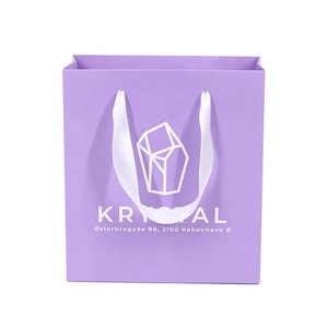 May include: A lavender paper shopping bag with white satin handles. The bag features the word "KRYSTAL" in white, along with a geometric logo and address text. The bag is square and has a simple, elegant design.