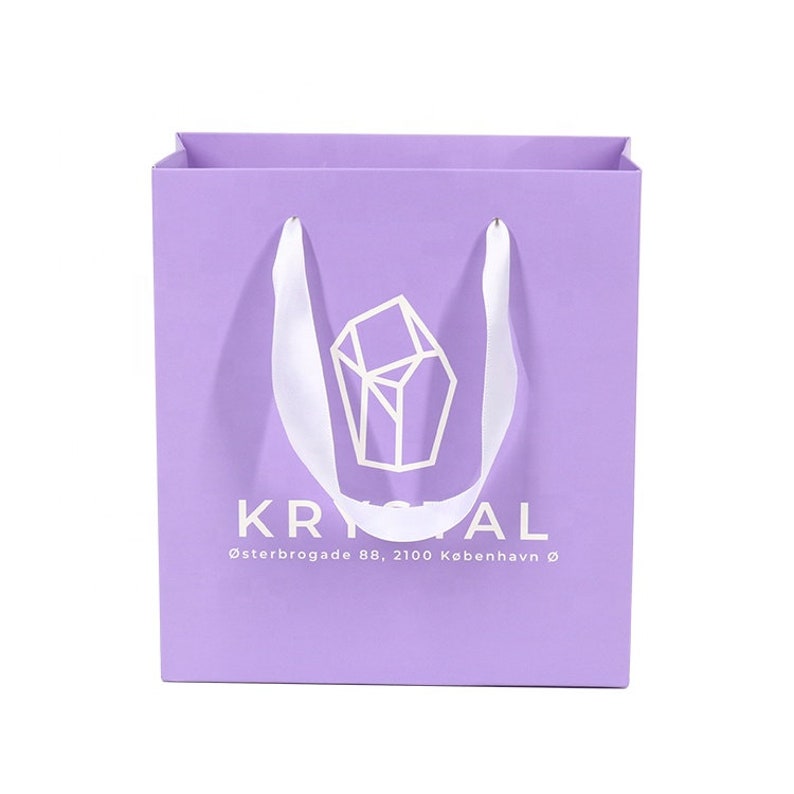 May include: A lavender paper shopping bag with white satin handles. The bag features the word "KRYSTAL" in white, along with a geometric logo and address text. The bag is square and has a simple, elegant design.